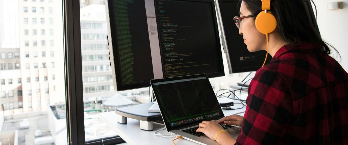 Woman programming on a laptop at a standing desk in an office with large windows.